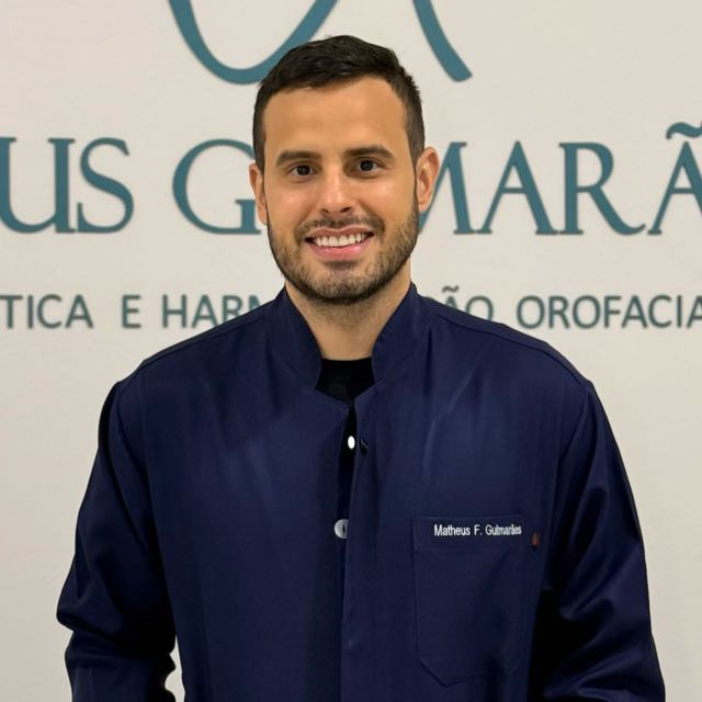 Dr. Matheus Guimarães sorrindo, usando jaleco azul-marinho